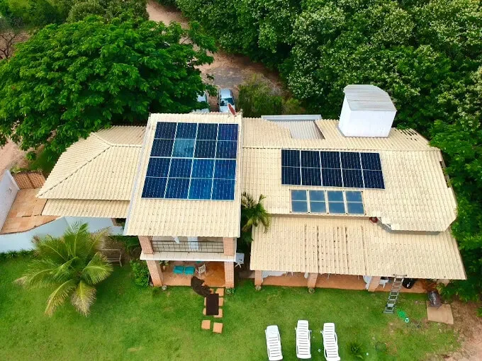 Casa de telha clara vista de cima, com dois conjuntos de módulos solares em águas diferentes do telhado e área verde em volta.