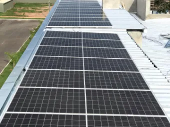 Telhado metálico coberto por módulos solares em fileiras contínuas, com área verde ao fundo.